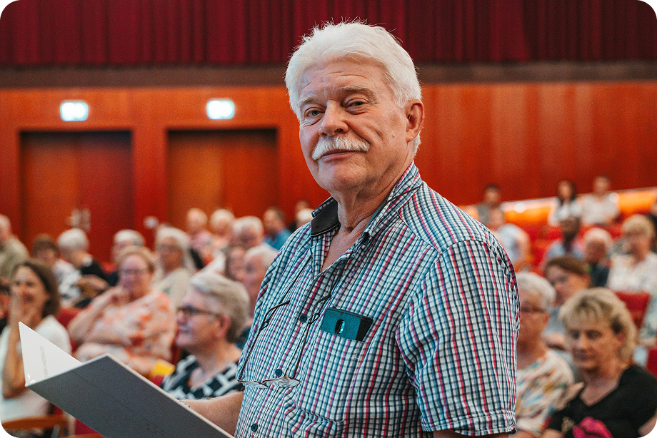 Ein älterer Mann schaut, stehend vor einer Gruppe auf der Mitgliederversammlung, in direkt die Kamera.
